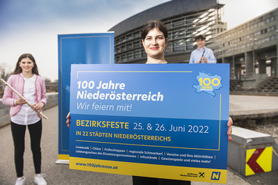 Eine Frau steht im Vordergrund mit einem Schild in der Hand, auf dem für die Bezirksfeste am 25. und 26. Juni geworben wird. Links davon steht eine junge Frau mit Querflöte und weiter im Hintergrund steht ein junger Mann mit Klarinettte. Das Foto wurde vor dem Landhaus St. Pölten aufgenommen.