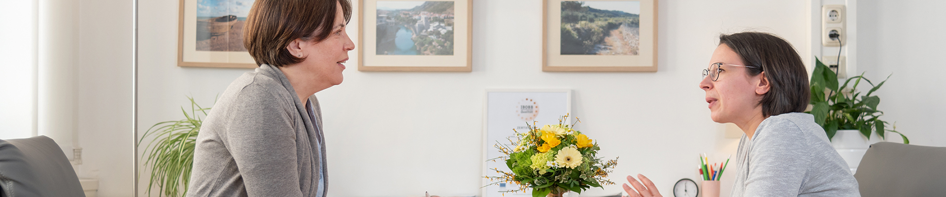 Zwei Frauen sitzen sich an einem runden Tisch in einem hellen Büro gegenüber und führen ein Beratungsgespräch. Im Hintergrund sind Bilder an der Wand, ein Blumenstrauß auf dem Tisch und Arbeitsmaterialien zu sehen, was eine ruhige, professionelle Atmosphäre vermittelt.