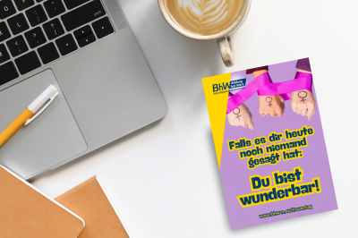 Schreibtisch von oben. Man sieht einen Laptop, einen Stift und eine Postkarte zum BhW Frauentag