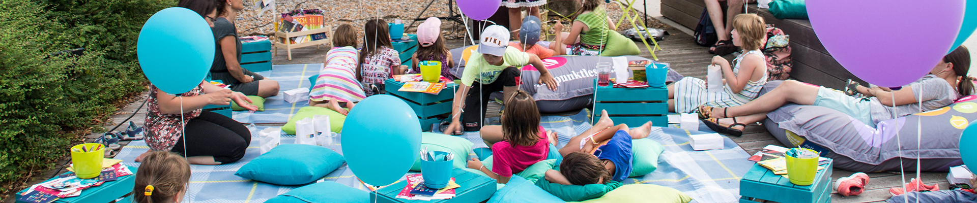 Kinder sitzen auf Decken und Pölter im Park und lauschen einer Geschichte.