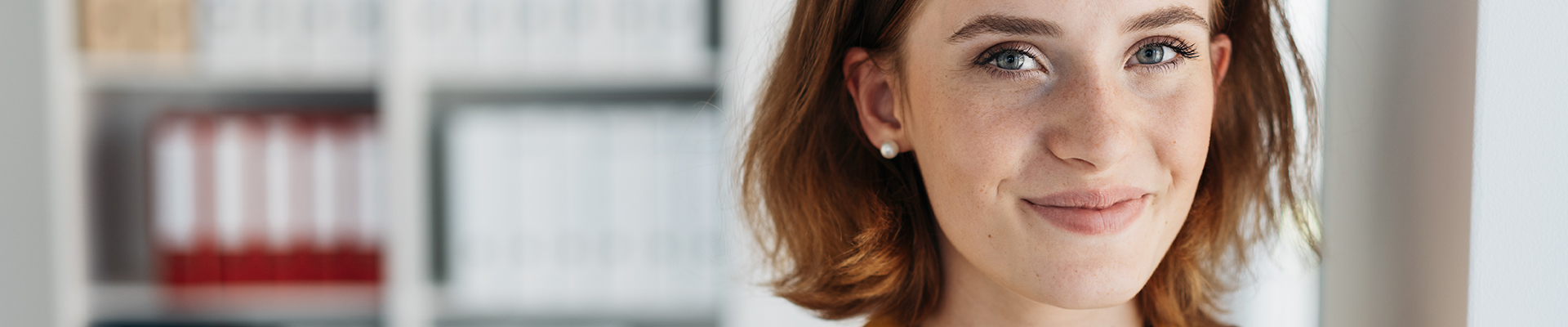 Portraitfoto einer jungen Frau mit roten Haaren. Sie trägt eine dottergelbe Bluse und lächelt in die Kamera. Im Hintergrund sieh tman ein Bücherregal.