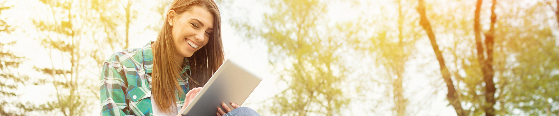 Das Headerbild zeigt eine junge Frau am linken Bildrand sitzend, die lächelnd und interessiert auf ihr Tablet sieht. Sie befindet sich im Freien, hinter ihr sind Bäume zu sehen.
