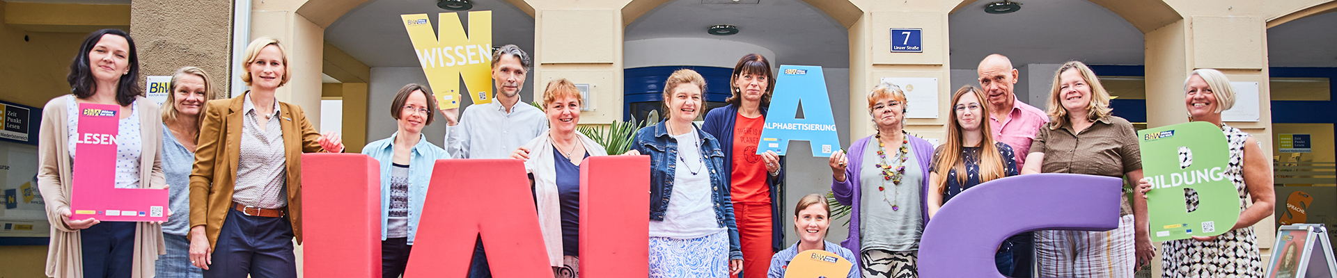 Die Lernbegleiterinnen und Lernbegleiter der Basisbildung stehen vor dem Haus Linzerstraße 7. Davor stehen noch zwei sehr große Buchstaben aus Schaumstoff. Ein großes "W" in rot und ein großes "S" in lila.  Einige Personen halten auch kleinere Buchstaben in den Händen.