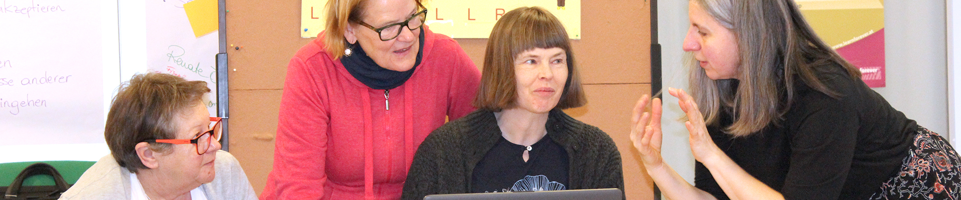 Eine ältere Dame sitzt am Tisch vor einem Computer. Rechts daneben sitzt noch eine Dame und dahinter steht eine Dame. Eine Lernbegleiterin steht links von ihnen und erklärt etwas.