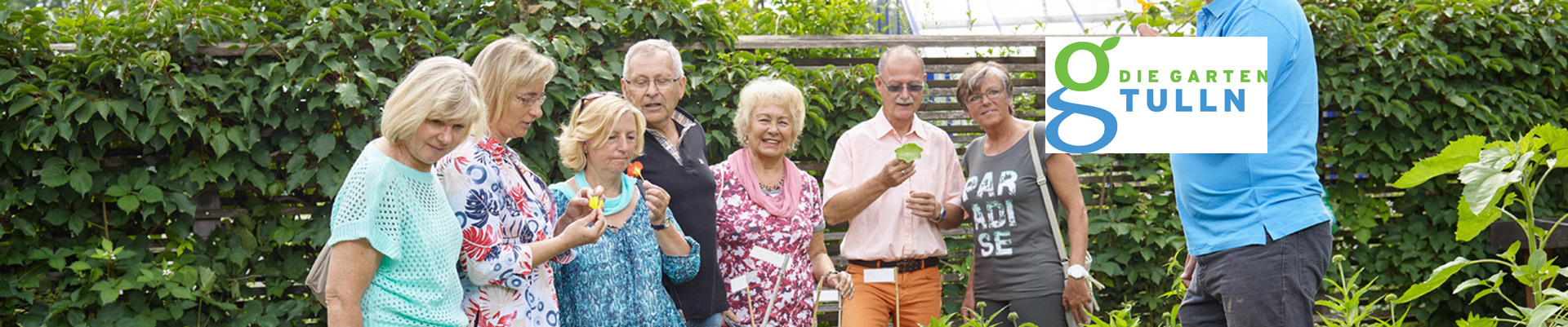 Das Headerbild zeigt eine Personengruppe, die bei einem Pflanzenbeet steht und von einem Gärtner essbare Blüten zum Verkosten bekommt.